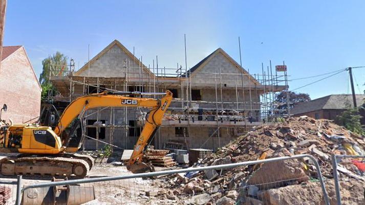 A JCB digger is pictured in front of a partially finished redevelopment site of a building. A large mound of rubble can be seen in front of the building which is surrounded in scaffolding. 