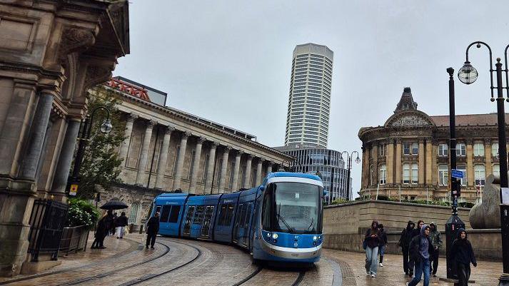 A blue and silver tram travels between stone buildings on a street with people wearing hoodies to protect from the rain, walking alongside.