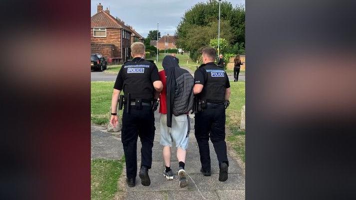 Two police officers dressed in black holding a man by his arms and leading him down a residential street. He's dressed in shorts and a red t-shirt and covering his head with a tracksuit top.