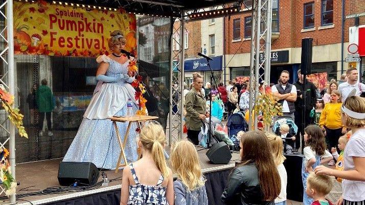 A stage with a woman dressed as Cinderella in a long blue gown and blonde wig with a bob singing into a microphone. Behind her is a orange and yellow sign saying Spalding Pumpkin Festival with decorations of autumn leaves. Children are stood at the front of the stage watching her perform and you can see a few high street shops to the side.