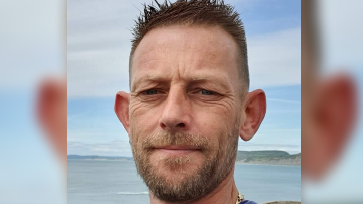 Stuart with short dark hair smiling at the camera with the sea behind him in the distance