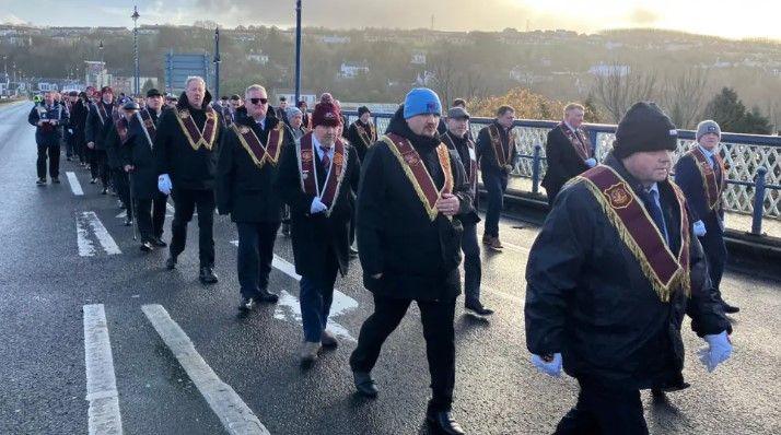 Apprentice Boys pictured in regalia, some wearing large coats and woolly hats, marching across the Craigavon Bridge.