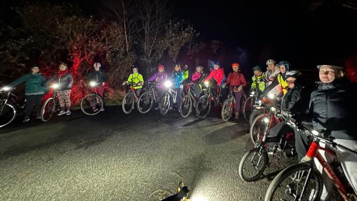 Fifteen cyclists are visible gathered in a semi circle on the road.
They are wearing helmets and their  bike lights are on.