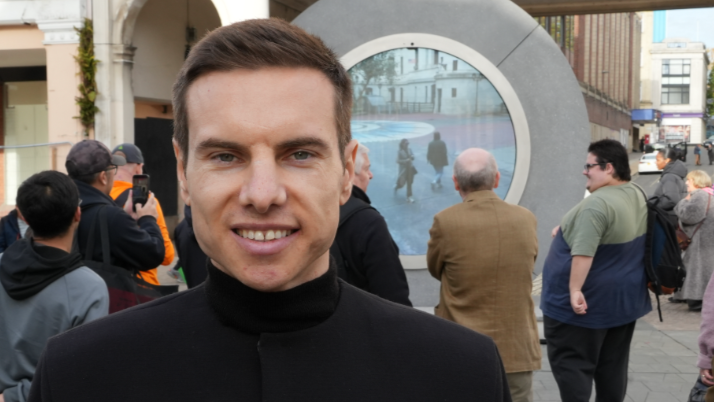 Benediktas Gylys was standing on the left of the image looking towards the camera smiling. He had short brown hair and was wearing a black turtle neck and coat. Behind him was a portal which was a circle made from stone with a screen in the middle.