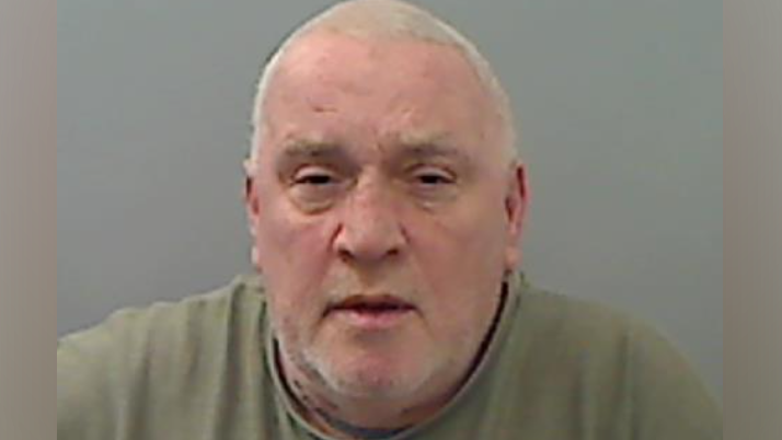 Edwin Youll with short white hair, wearing a green top, looks directly at the camera in a police mugshot photo, with a grey background.
