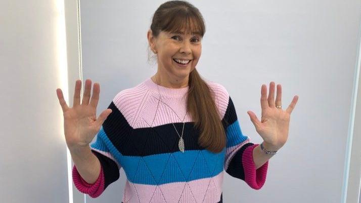 Karen Dingle wears a jumper with pink, black and azure blue horizontal stripes. She is smiling at the camera, her long brown hair is tied in a pony over her left shoulder and she is standing in front of a white wall.