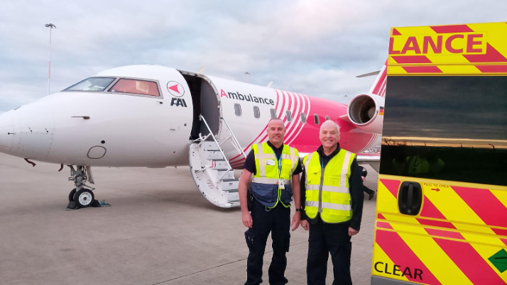 Two ambulance men, wearing bright green jackets, smile in front of the medical plane