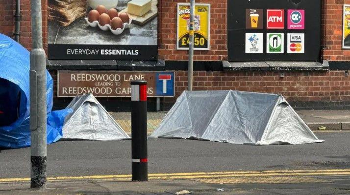 Two silver small tents and blue tarpaulin over a car next to the Reedswood Lane sign