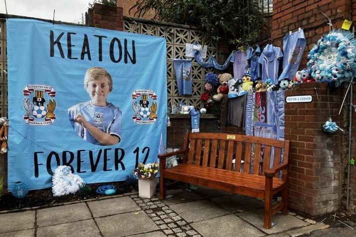 A corner of a garden with lots of tributes, most of them blue. A large light blue poster with Keaton in his Coventry City kit on reads 'Keaton Forever 12', with two Coventry City logos on it. Several Coventry City shirts are hung up and there is a sign reading 'Keaton's Corner'.