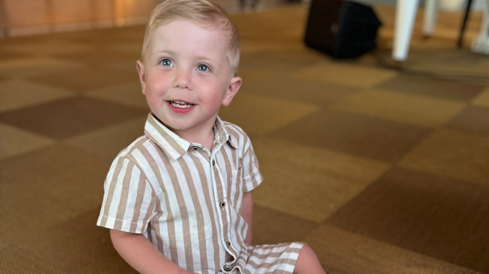 Hudson is wearing a brown and white stripy top and shorts and is sitting on the floor smiling at the camera