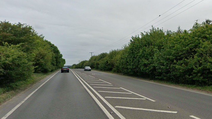 A general view of the A143 at Ixworth. Vehicles travel either side of the road. The road is lined with trees.