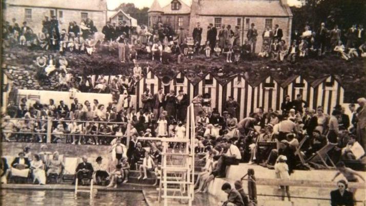 A black and white photo donated to the pool shows a very busy sea pool with changing rooms that look like beach huts and families crowded around the water sitting in deckchairs.