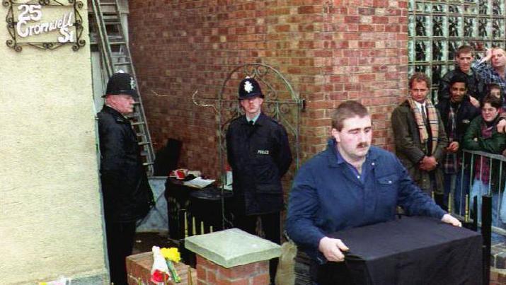 A man carrying a black box out of the house in Cromwell Street where Fred and Rose West lived. A crowd of people and two police officers are watching on.