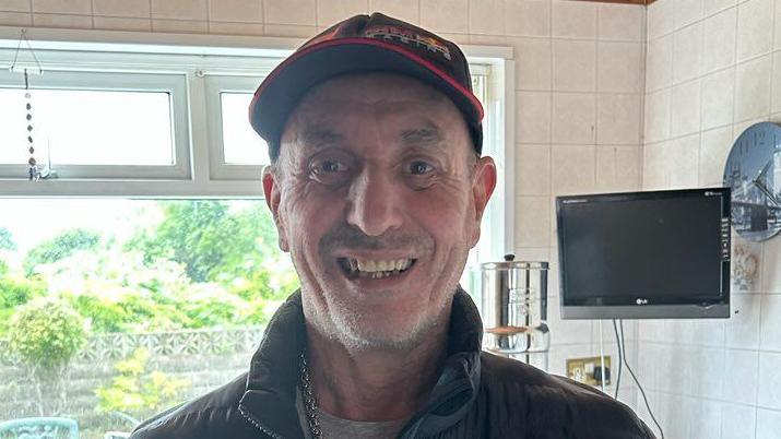 Llun o Byron Long. Mae'n gwisgo cap du a choch, crys-t llwyd a chôt ddu. Mae'n sefyll mewn cegin sy'n edrych allan ar ardd. Mae teledu a chloc ar y wal y tu ôl iddo hefyd.