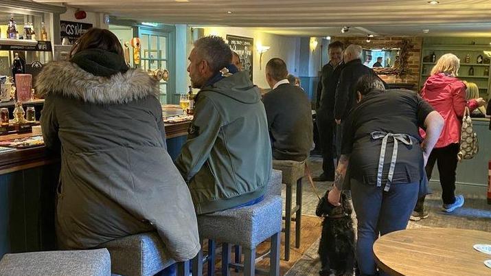 The interior of a busy country pub with three people on stools sitting at the bar and a member of a staff stroking a black spaniel. Behind her there are more people sitting and tables and chatting 