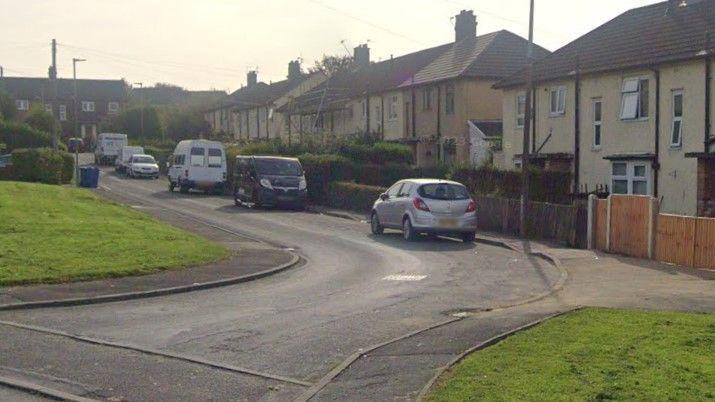 Google Earth photograph of Pilmuir Road in Blackburn. Cars and vans are parked along the road, with a row of semi-detached houses and more homes at the top of the road. Two grass verges can be seen on either side of the entrance to the road.
