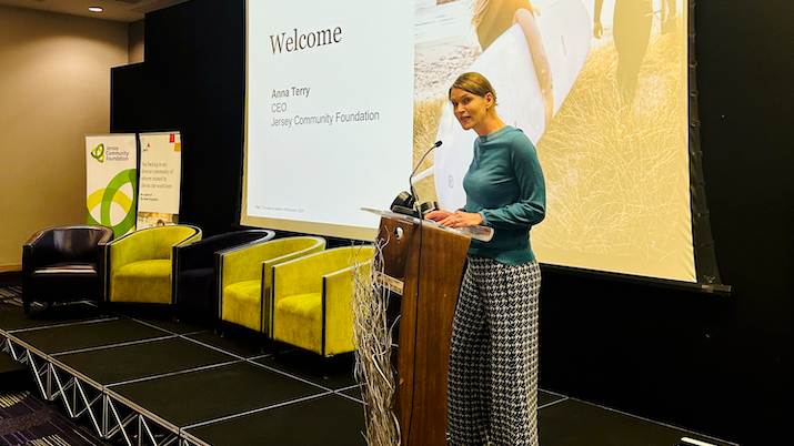 A woman standing on stage behind a podium speaking into a microphone with a powerpoint behind showing a welcome screen. She is wearing a long sleeve blue top a checkered black and white wide trousers.