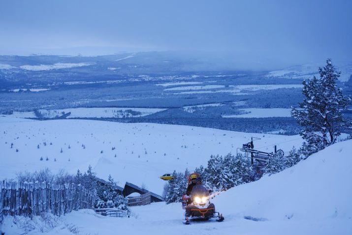 A person rides a snow mobile up a snow-covered slope in the Cairngorms. There is a snow covered landscape sprawled out behind the rider, a patchwork of fields and forestry.