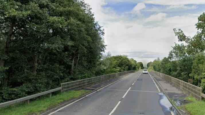 A road over a bridge, lined with crash barriers. There is greenery and trees on either side of the road, with a white car approaching the camera.