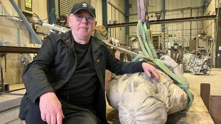 A man with a black hat, glasses, a black jacket, black shirt and jeans. He is sat down on a work table in a workshop and has his left hand on a statue of Lemmy, which is lying down.