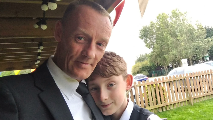 Lee Kervin is photographed with his son Brad, who is in his mid-teens in the photograph. Lee is wearing a black suit, white shirt and black tie, while Brad is wearing a white shirt and black waistcoat. They are outdoors, under a trellis, with some grass and a fence in the background. 