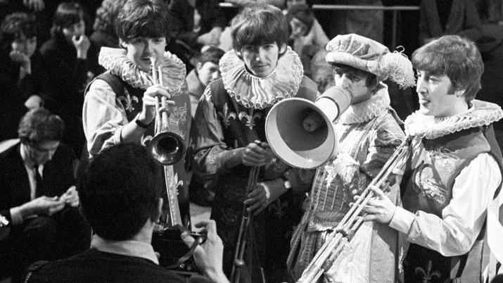 The Beatles, dressed as page boys from the Middle Ages, are photographed holding brass instruments.