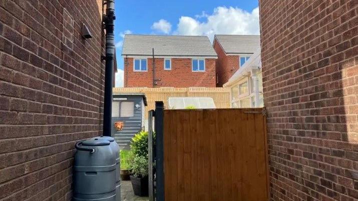 A gap in between two red brick houses, with a row of houses in the background. There is a fence panel and two water butts in the foreground. 