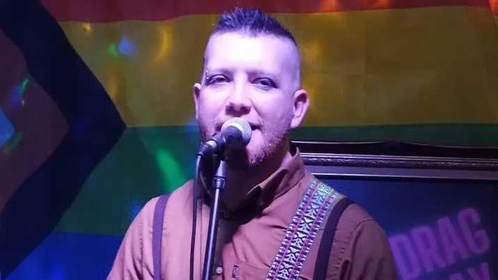 Patrick appears to be singing into a microphone, with blue lighting behind him. He has short spikey hair and a piercing in one eyebrow. He wears a brown shirt. He has a guitar strap over his shoulder.