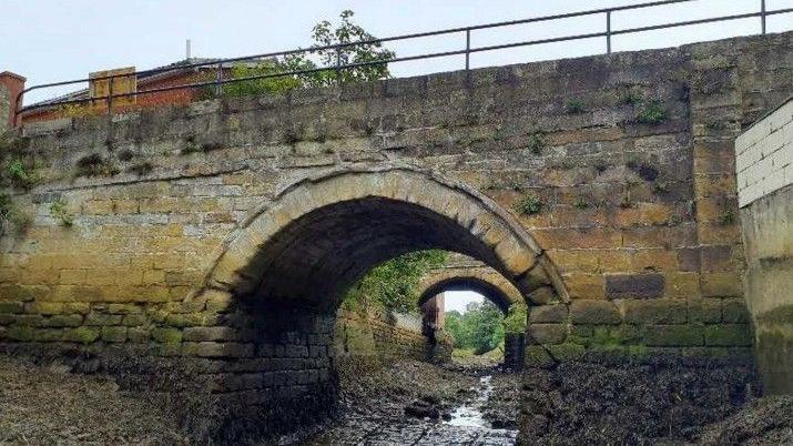 Spital Old Bridge, a traditional stone bridge over a mostly dry beck, with water marks running up each side.