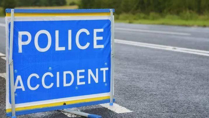 A blue sign on the side of a road which says 'Police Accident'. Bushes can be seen on the other side of the road, which is clear. 