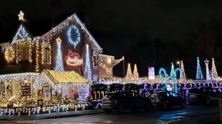 A house is covered with festive lights and there are tall illuminated structures in the garden which are shaped like cones. The lights are different colours, and cover most parts of the building and surrounding fence and wall.