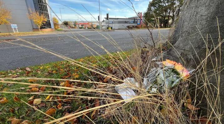 Yellow roses left at the foot of a tree on a verge by a road. In the background grey buildings are visible on what appears to be a retail estate.