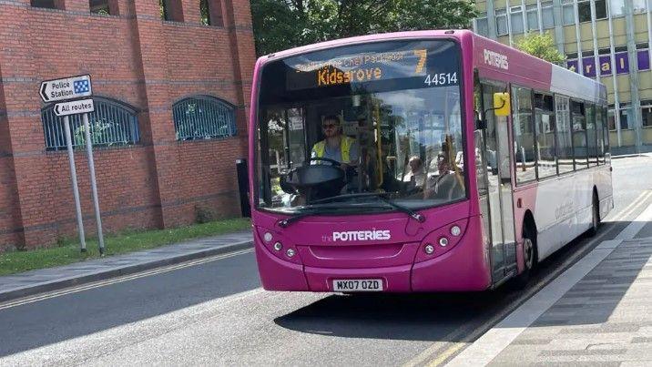 A purple and white Potteries bus which says Kidsgrove on the display above the windscreen. It is being driven along a road with the driver visible through the window.
