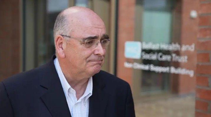 Ciaran Mulgrew is looking to the right of the frame. He is wearing thin framed glasses and has a plain expression. He is bald and wearing a navy suit jacket, and a white shirt with no tie and the top button undone.