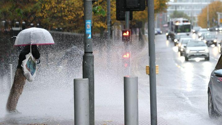 A pedestrian is standing on the side of a busy road in heavy rain and a stream of cars drive past through a large puddle. They are wearing a black jacket, brown trousers and are holding a clear plastic umbrella.