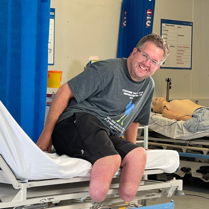 Neil Hopper is sitting on the side of a hospital bed wearing a grey T-shirt, black shorts and glasses. He is not wearing prosthetic limbs so his bilateral leg amputations are on show. He is smiling at the camera.
