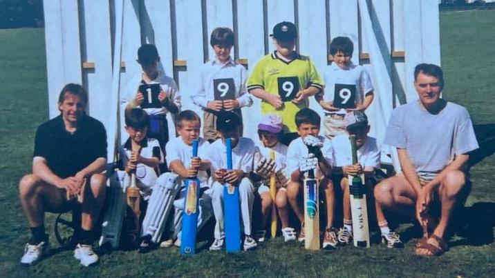 Ten children, some wearing caps and holding bats and one wearing pads, in a team line up in front of a white sight screen. A man sits to the left of the front row and a second man to the right. Four children who holding individual numbers which make the year 1999.