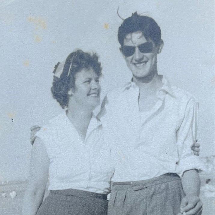 David Balfour with his mother Doris in 1957. Both are smiling and wearing white shirts. He is wearing sunglass and has his arm around his mother. The sea can be seen in the background.