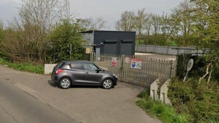 Grey prefab buildings with roller shutters in a plot alongside a rural road. The site is surrounded by trees and there are metal gates across the entrance, which have security company warning notices on them. A small grey car is parked at the entrance.