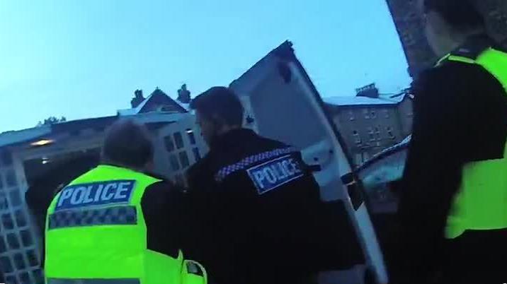 Body-worn camera footage of two police officers in hi-vis vests and one in a black police vest, at the back of a police van. The police van back doors are open. Snow-covered roofs of residential buildings can be seen behind the van.