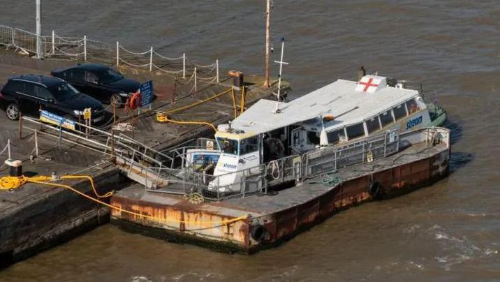 Aerial view of a grey ferry docked next to a car park. The water is brown