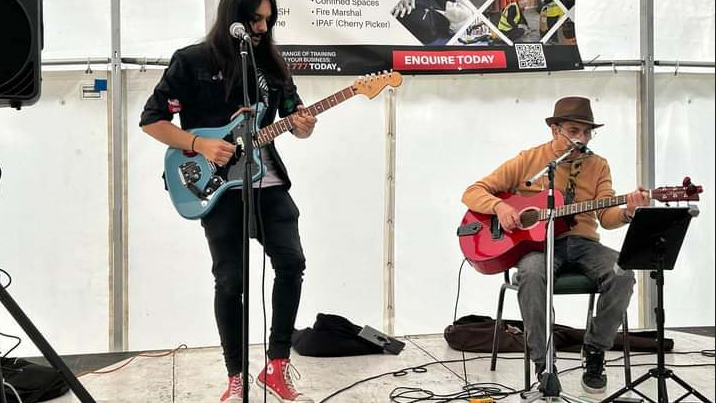 Bansal Electrics are two people with one pictured standing playing a guitar and the other seated with a guitar. The man seated is wearing a brown jacket and grey jeans and the other band member is all in black with red trainers.