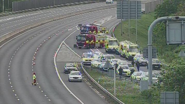 A CCTV screenshot showing two fire engines and a several ambulances and a police car lined up on a motorway slip road.