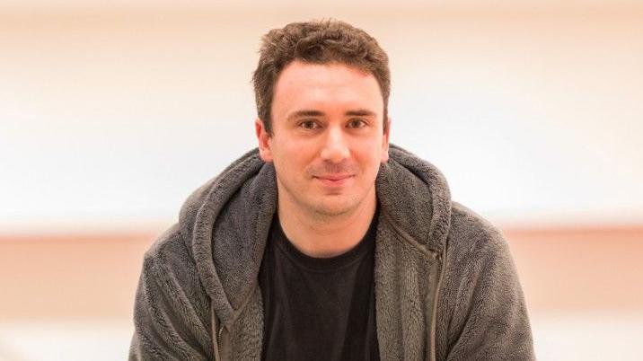 Sam Smith is looking at the camera. He is wearing a grey fleece hoodie and black tee shirt and has short brown hair.