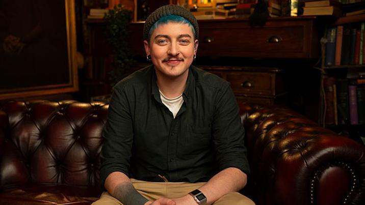 Alex, with blue hair and a beanie hat, sits on a brown leather Chesterfield sofa, smiling. He is wearing chinos and a jumper and white T-shirt.