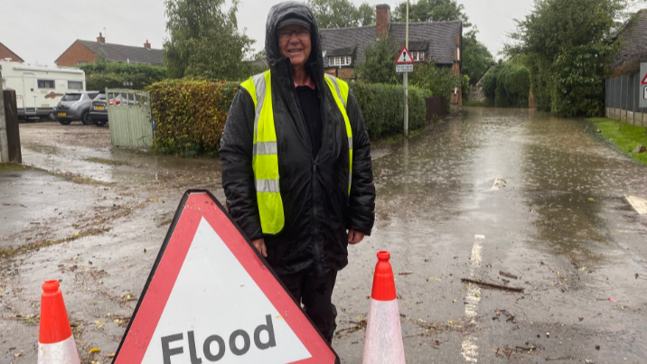 A man in waterproof clothing, wearing a hi-vis vest, stands next to a triangular flood warning sign and two traffic cones.  Rain falls and the road behind him is submerged by water 