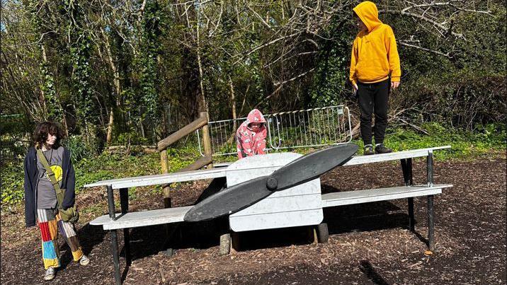 Three children play on play park equipment shaped like an aeroplane. One child stands on the left and is wearing colourful trousers, a grey top and a black zip up hoodie. The child in the middle wears a pink onesie and is crouched on top of the aeroplane. And the third stands on the left propeller and wears a yellow hoodie and black trousers.