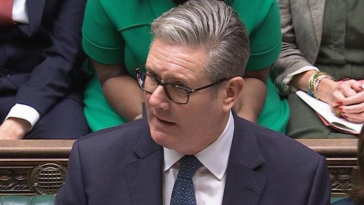 Sir Keir Starmer  addresses MPs in the House of Commons. people site in the pews and behind him one can see the hands and forsos of two females and a man. 