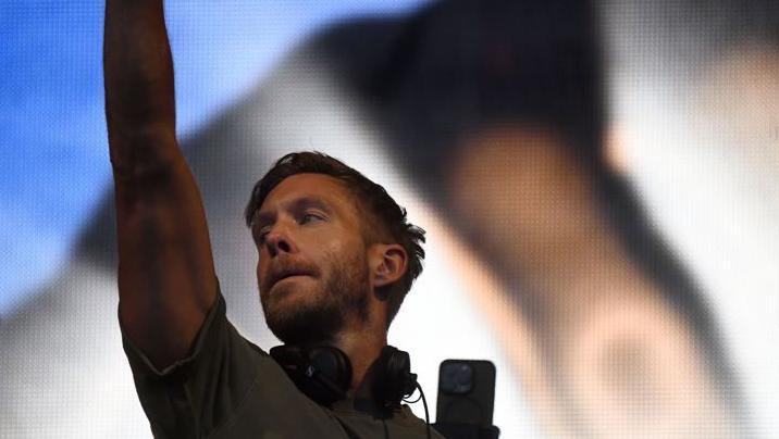 Calvin Harris wearing headphones around his neck raises his arm in the air on stage 