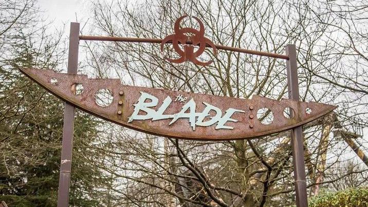 The signage above the ride entrance, with trees behind. It is a stylised rusty industrial blade attached to two poles, with a bar above it. The blade has lettering on it which reads The Blade.